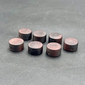 Könnte beinhalten: Sieben kleine, zylindrische Holzknebel in dunklem Rotbraun. Jeder Knebel hat eine flache Ober- und Unterseite mit einem kleinen Loch, das durch die Seite gebohrt wurde. Die Knöpfe sind auf einer dunkelgrauen Oberfläche angeordnet.