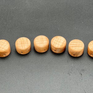Peut inclure: Six petites perles cylindriques en bois clair, présentant un motif de grain visible. Chaque perle est percée d'un petit trou central, adapté à l'enfilage. Les perles sont disposées en ligne.