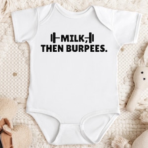 May include: White baby onesie with the text "MILK, THEN BURPEES." in black. The text is accompanied by a barbell graphic. The onesie is short-sleeved and has a simple, classic design.