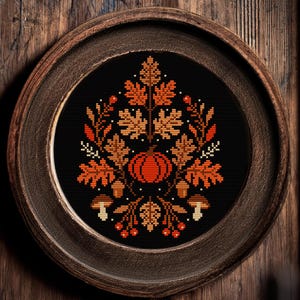 May include: A framed cross-stitch featuring a fall-themed design. The artwork showcases a central pumpkin surrounded by leaves, berries, and mushrooms in shades of orange, brown, and beige against a black background. The round wooden frame adds a rustic touch.