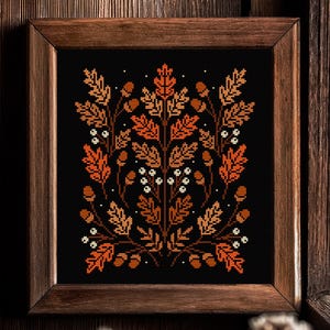 Könnte beinhalten: Kreuzstich-Kunstwerk mit einem symmetrischen Design aus Herbstblättern, Eicheln und Beeren in Orange-, Braun- und Weißtönen auf schwarzem Hintergrund. Das Kunstwerk ist in einem dunkelbraunen Holzrahmen gerahmt.