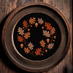 May include: A framed cross-stitch of an autumn wreath. The wreath features oak leaves and acorns in shades of orange and tan against a black background. The round frame is made of dark brown wood, and the background is a wooden surface.