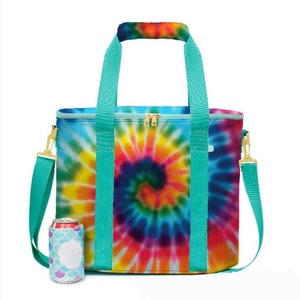 May include: A tie-dye insulated cooler bag with a spiral design in vibrant colors. The bag has teal handles, a shoulder strap, and gold-colored hardware. A matching can cooler with a mermaid scale pattern is next to the bag.