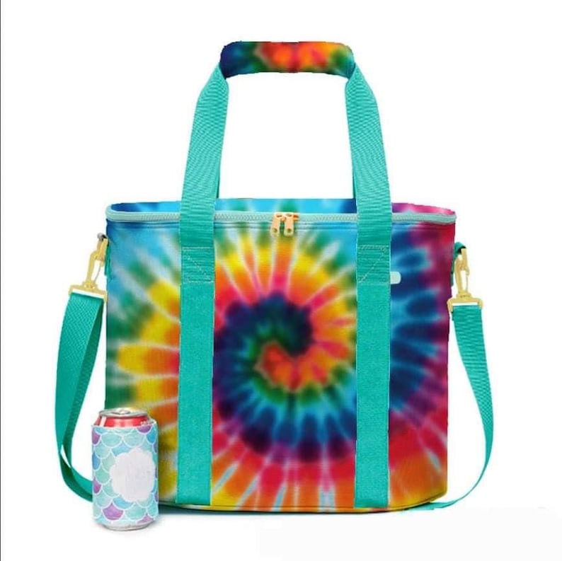 May include: A tie-dye insulated cooler bag with a spiral design in vibrant colors. The bag has teal handles, a shoulder strap, and gold-colored hardware. A matching can cooler with a mermaid scale pattern is next to the bag.
