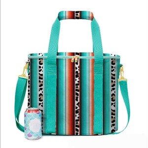 May include: A turquoise and brown striped cooler bag with a matching handle and shoulder strap. The bag features a gold zipper and a leopard print accent. A can cooler with a mermaid scale design is next to the bag.