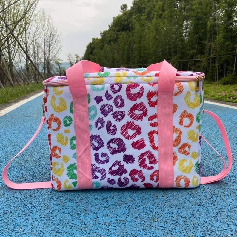 May include: A white cooler bag with a colorful leopard print design. The bag features pink handles and a shoulder strap. The leopard print includes red, purple, yellow, green, and orange spots. The bag is designed for carrying food and drinks.