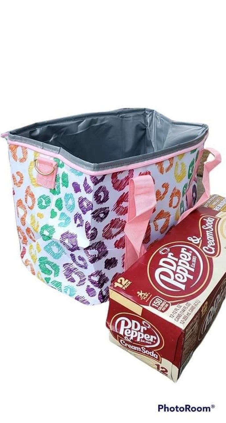 May include: A colorful tote bag with a rainbow leopard print design, pink handles, and a gray interior. Next to the bag is a 12-pack of Dr. Pepper & Cream Soda.