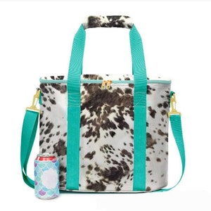 May include: A rectangular cooler bag with a cowhide pattern in black and white. It has teal handles and a teal shoulder strap with gold-colored hardware. A can with a mermaid-themed koozie sits next to the bag.