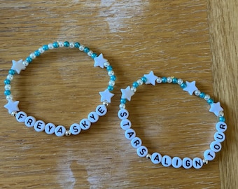 May include: Two beaded bracelets with white star-shaped beads and turquoise and pearl-coloured beads. One bracelet spells "FREYA SKYE" and the other spells "STARS ALIGN TOUR" in black letters on white beads. The bracelets are on a light brown surface.