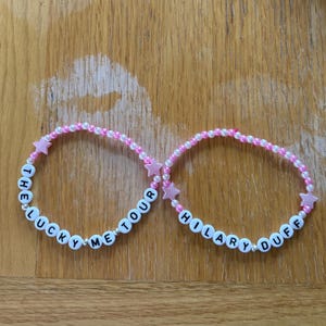 May include: Two beaded bracelets with white letter beads spelling "THE LUCKY ME TOUR" and "HILARY DUFF". The bracelets have pink star and pearl accents. The background is a light brown wooden surface.
