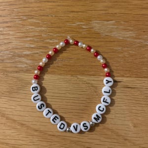 May include: A beaded bracelet with white letter beads spelling "BUSTED VS MCFly". The bracelet features alternating red and cream-coloured beads. The bracelet is on a wooden surface.