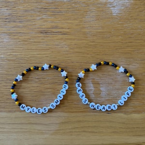 May include: Two beaded bracelets. One spells "MYLES SMITH", the other "MESS HEART LIFE". The bracelets feature black, gold, and blue beads, with white star-shaped beads. The bracelets are made of small beads.