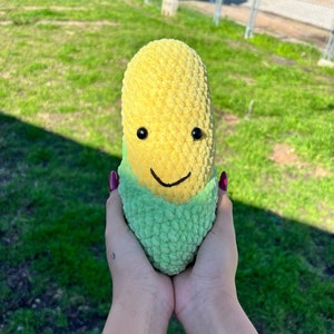 Könnte beinhalten: Ein Plüsch-Maiskolben-Spielzeug mit einem lächelnden Gesicht, grünem Hüllblatt und gelben Körnern. Das Spielzeug wird von zwei Händen gehalten.
