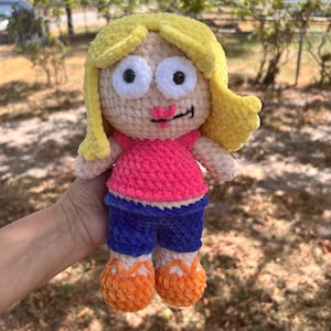 Puede incluir: Una muñeca de ganchillo hecha a mano con pelo de lana amarillo brillante, ojos grandes blancos con pupilas negras y una pequeña boca rosa bordada. La muñeca viste una camiseta rosa vibrante de manga corta, pantalones cortos azul oscuro y zapatos naranjas.