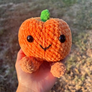May include: A plush orange pumpkin with a green stem and black button eyes. The pumpkin has a smiling face and small orange feet. The pumpkin is a handmade item.
