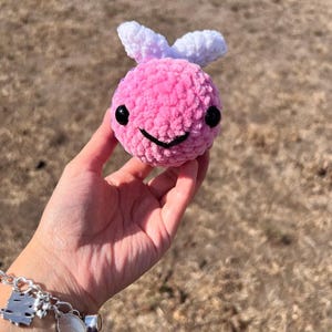 May include: A round, pink, plush crochet bee toy with a soft, fuzzy texture. It features two small white wings on top, two large black plastic safety eyes, and a simple black stitched mouth. The toy has a spherical body shape and is crafted from yarn.