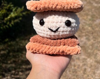 Könnte beinhalten: Ein handgefertigtes Häkel-S'more-Spielzeug mit einem lächelnden Gesicht. Das S'more hat eine hellbeige Marshmallow-Mitte und zwei Schichten aus braunen Graham-Crackern sowie eine obere Schicht aus braunen Graham-Crackern. Das Spielzeug wird in einer Hand gehalten.