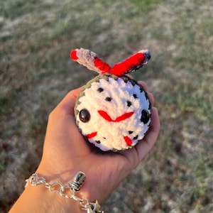 May include: A handmade, crocheted Jason Voorhees-inspired plushie. The round plushie features a white mask with black spots, a red mouth, and black eyes. It has red and gray accents on top. The plushie is held in a hand.