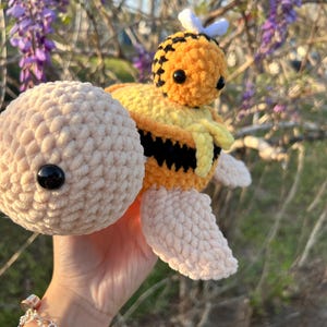 May include: A crocheted plush toy turtle with a crocheted bee on its back. The turtle is light pink and the bee is yellow and black with white wings.