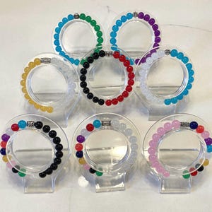 May include: A collection of beaded bracelets in various colors, including green, blue, red, black, white, yellow, and purple. Each bracelet is displayed on a clear, circular stand. The beads appear to be smooth and spherical.