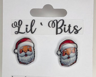 May include: A pair of Santa Claus earrings with a white background. The earrings are made of clear plastic with a printed image of Santa Claus's face. The earrings are packaged on a white card with the text "Lil' Bits" and "handmade in Michigan".