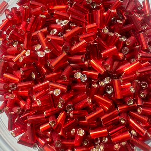 May include: A close-up view of a bowl filled with red glass beads. The beads are cylindrical in shape and have a shiny, metallic finish.