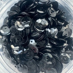 May include: A close-up view of a container filled with black sequins. The sequins are round and have a slightly raised center.