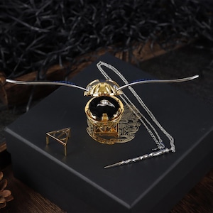 Könnte beinhalten: Eine goldfarbene Ringbox in Form eines goldenen Schnitzers aus der Harry-Potter-Reihe. Die Box ist geöffnet und ein Ring befindet sich darin. Die Box steht auf einer schwarzen Box mit einer silbernen Halskette und einem silbernen Zauberstab.