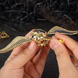 Puede incluir: Réplica dorada de la Snitch con alas negras y doradas, un objeto de colección de la serie Harry Potter. La Snitch es sostenida por manos, con el símbolo de las Reliquias de la Muerte en el fondo.