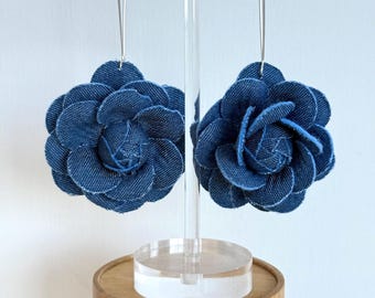 May include: A pair of denim flower earrings. Each earring features a large, layered blue denim flower suspended from a silver-colored hoop. The earrings are displayed on a clear acrylic stand with a wooden base, set against a white background.