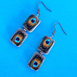 May include: A pair of dangle earrings featuring a rectangular design with a peacock feather pattern. The earrings have a blue, gold, and black color scheme, with silver-colored ear wires and connecting beads. The background is a solid blue.