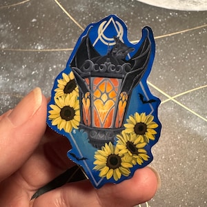 May include: A sticker featuring a black lantern with bat wings and a bat perched on top. The lantern has an orange glow and is surrounded by yellow sunflowers. The background is blue with a geometric design.