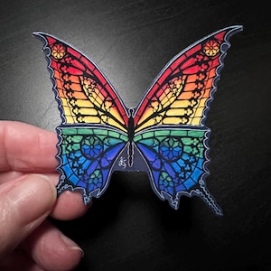 May include: A colorful butterfly sticker with stained glass wing designs. The wings transition from red to yellow to green to blue. The butterfly is held by a person's hand against a dark background.