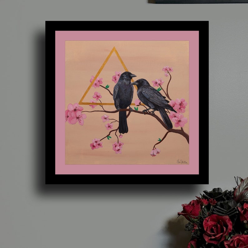 Black frame with a pink matte. Painting of two black crows on a cherry blossom branch and a unique golden metallic triangle.