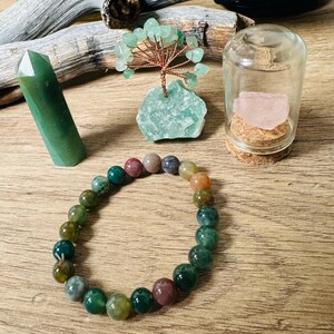 Puede incluir: Un brazalete de ágata verde con una variedad de piedras verdes, marrones y rosas. El brazalete está sobre una superficie de madera con un pequeño árbol de cristal verde, un frasco de vidrio con un cristal rosa y una punta de cristal verde.