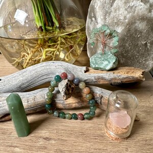 May include: A green agate bracelet with red, brown, and green beads. The bracelet is lying on a wooden surface next to a green crystal point, a small glass jar with a pink crystal inside, and a green crystal tree.