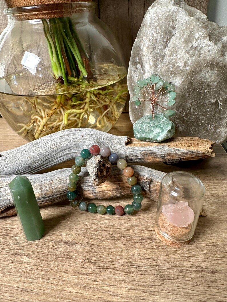May include: A green agate bracelet with red, brown, and green beads. The bracelet is lying on a wooden surface next to a green crystal point, a small glass jar with a pink crystal inside, and a green crystal tree.