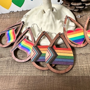 May include: Set of six rainbow-themed wooden earrings. The earrings are shaped like teardrops and feature a rainbow flag, a heart, and a chevron pattern.