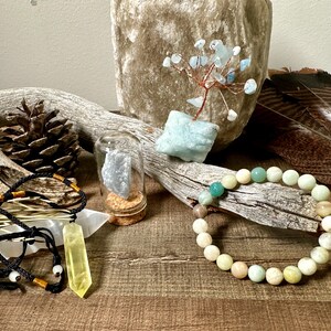 May include: A collection of crystals and stones, including a green and white beaded bracelet, a yellow crystal pendant on a black cord, a clear glass jar with a blue crystal inside, and a small tree made of wire and blue crystals.