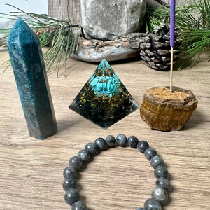 May include: A crystal pyramid with a turquoise stone inside, a dark blue crystal point, a grey beaded bracelet, and a wooden incense holder with a purple incense stick.