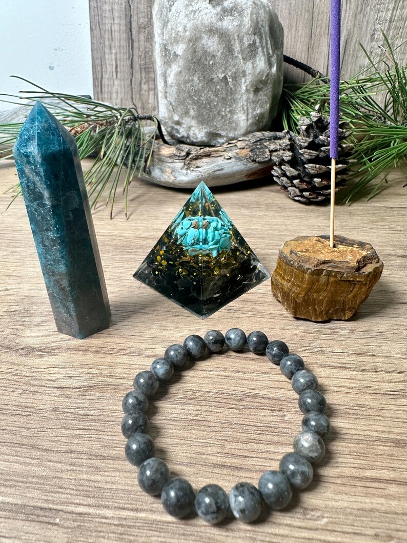 May include: A crystal pyramid with a turquoise stone inside, a dark blue crystal point, a grey beaded bracelet, and a wooden incense holder with a purple incense stick.