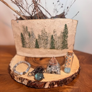 Puede incluir: Un conjunto de cristales y joyas sobre una rodaja de madera. El conjunto incluye una botella de vidrio transparente con un tapón de corcho llena de cristales azules, un cristal gris en forma de pirámide, un collar de cordón marrón con un colgante de labradorita gris y azul, y una pulsera de cuentas grises.