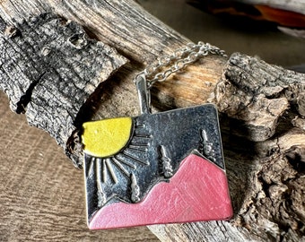 May include: A silver pendant necklace with a square charm featuring a pink mountain range, a yellow sun, and silver trees.