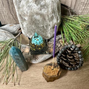 May include: A purple incense stick is burning in a gold and brown stone incense holder. A teal blue crystal, a pyramid-shaped crystal with turquoise and gold accents, and a grey Himalayan salt lamp are also in the image.