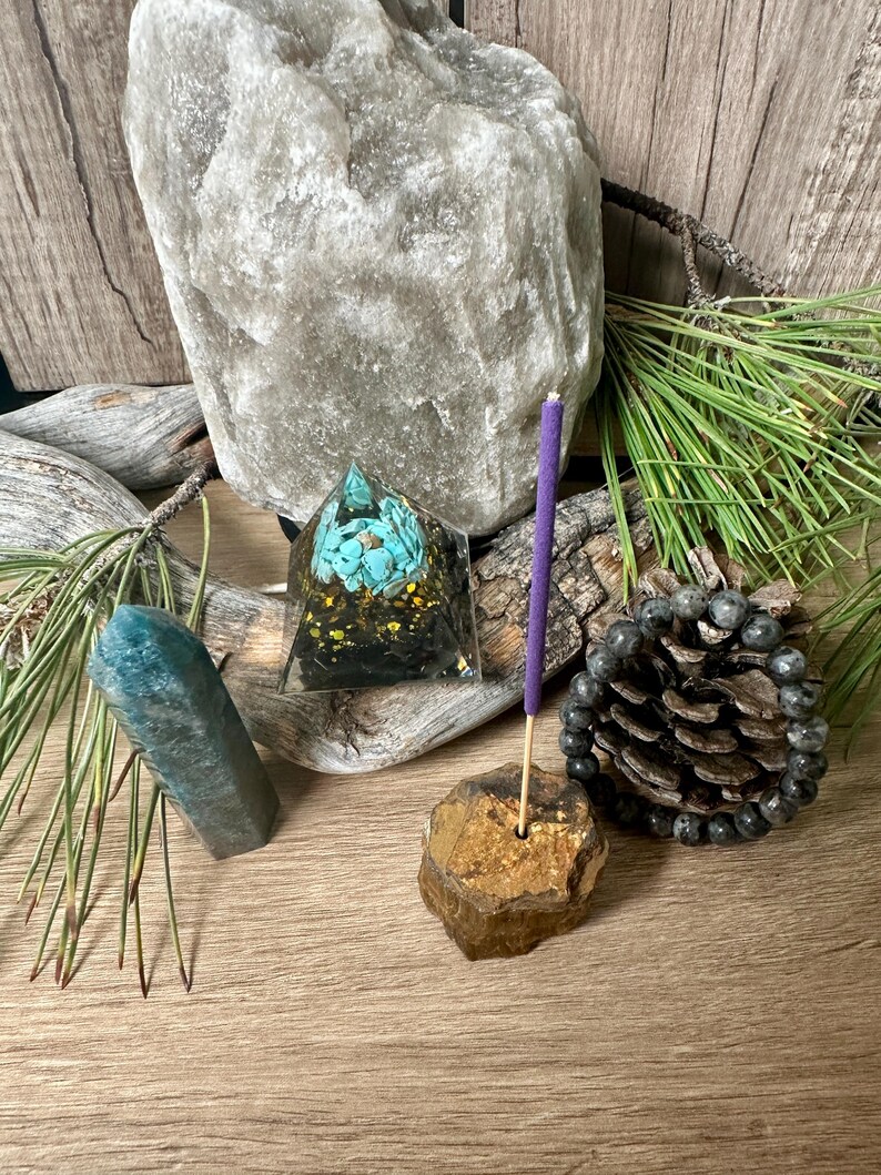May include: A purple incense stick is burning in a gold and brown stone incense holder. A teal blue crystal, a pyramid-shaped crystal with turquoise and gold accents, and a grey Himalayan salt lamp are also in the image.