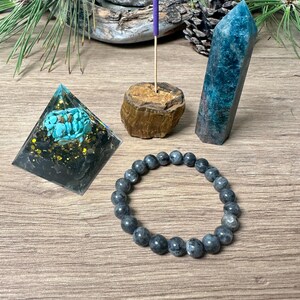 May include: A crystal pyramid with turquoise and gold accents, a blue-green crystal point, a wooden incense holder with a purple incense stick, and a grey beaded bracelet.