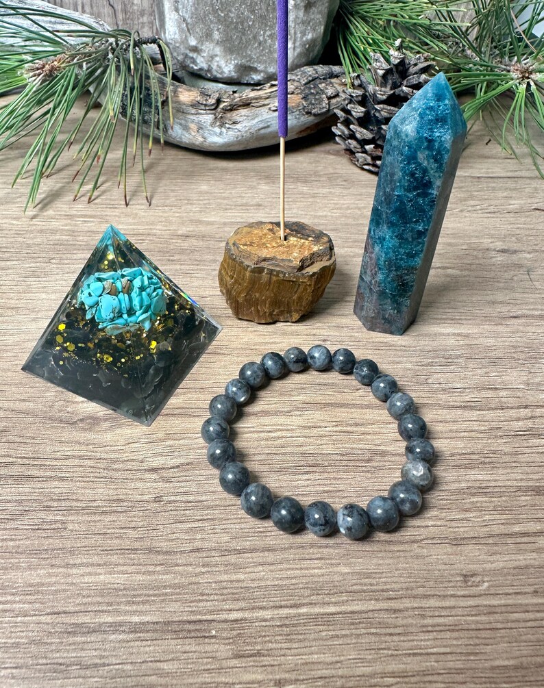 May include: A crystal pyramid with turquoise and gold accents, a blue-green crystal point, a wooden incense holder with a purple incense stick, and a grey beaded bracelet.