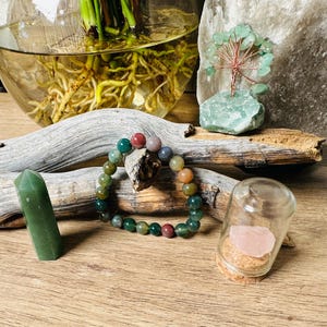May include: A multi-colored agate gemstone bracelet with a variety of green, red, and brown beads. The bracelet is displayed on a wooden surface with a small glass jar containing a pink crystal and a green crystal point.