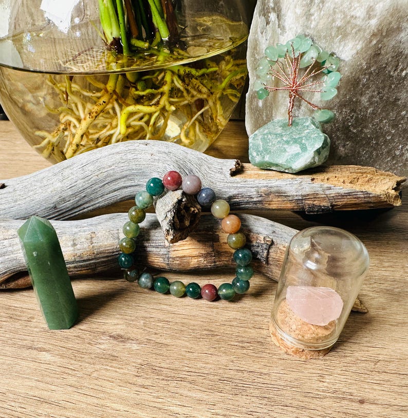 May include: A multi-colored agate gemstone bracelet with a variety of green, red, and brown beads. The bracelet is displayed on a wooden surface with a small glass jar containing a pink crystal and a green crystal point.