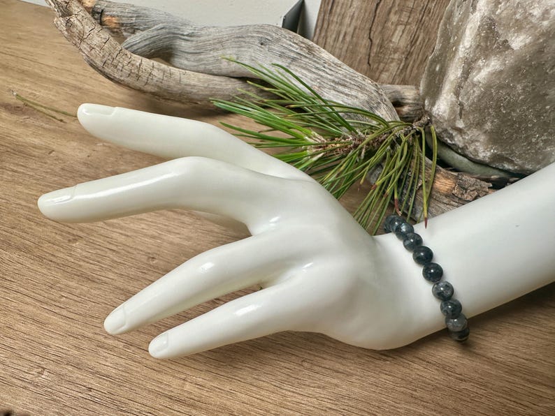 May include: A black beaded bracelet with dark grey and silver flecks. The bracelet is made of round beads and is worn on a white mannequin hand.